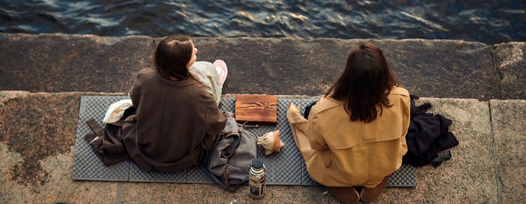 two women bundled up, sit next to the water on mats